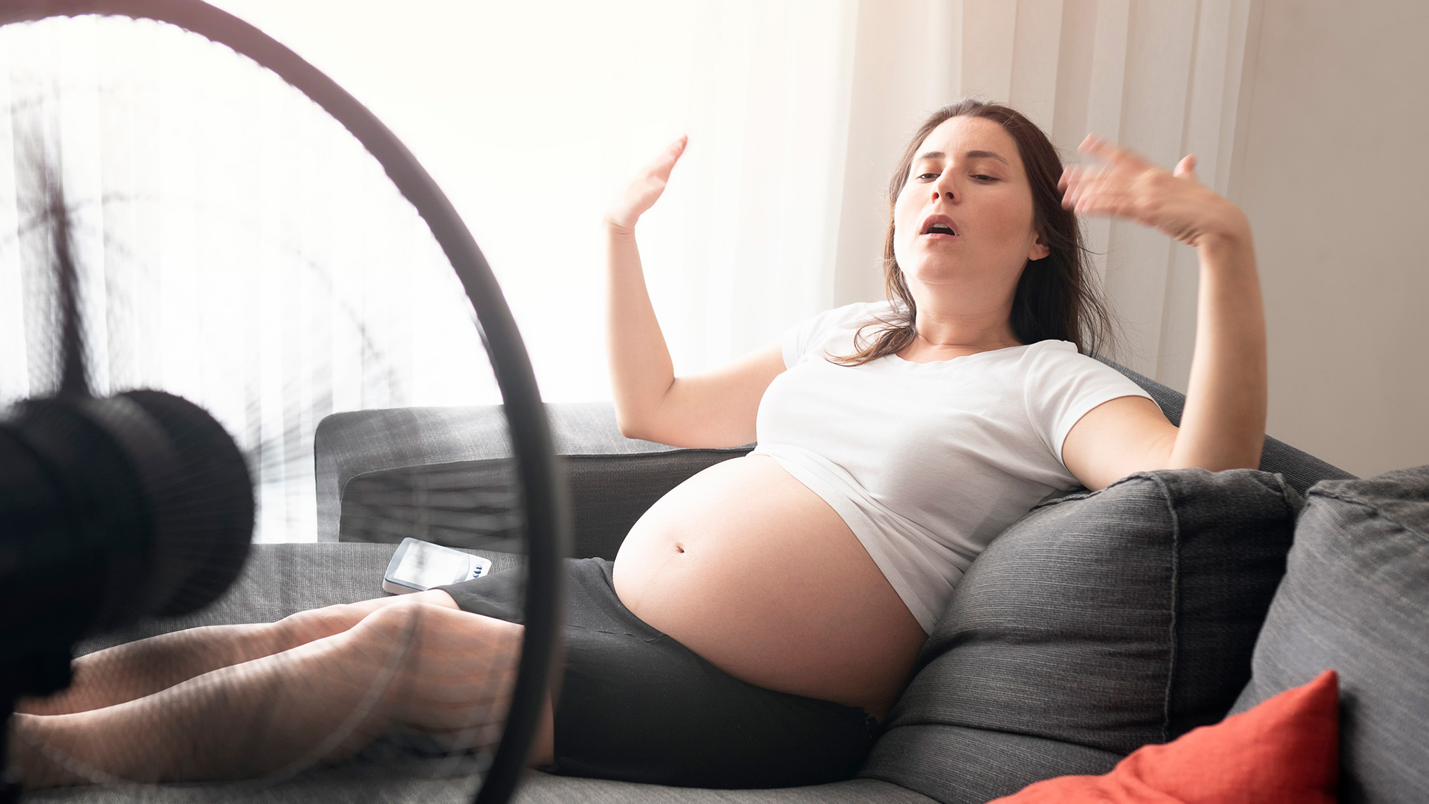 A pregnant woman wearing a white t-shirt fans herself while laying in front of a large fan.