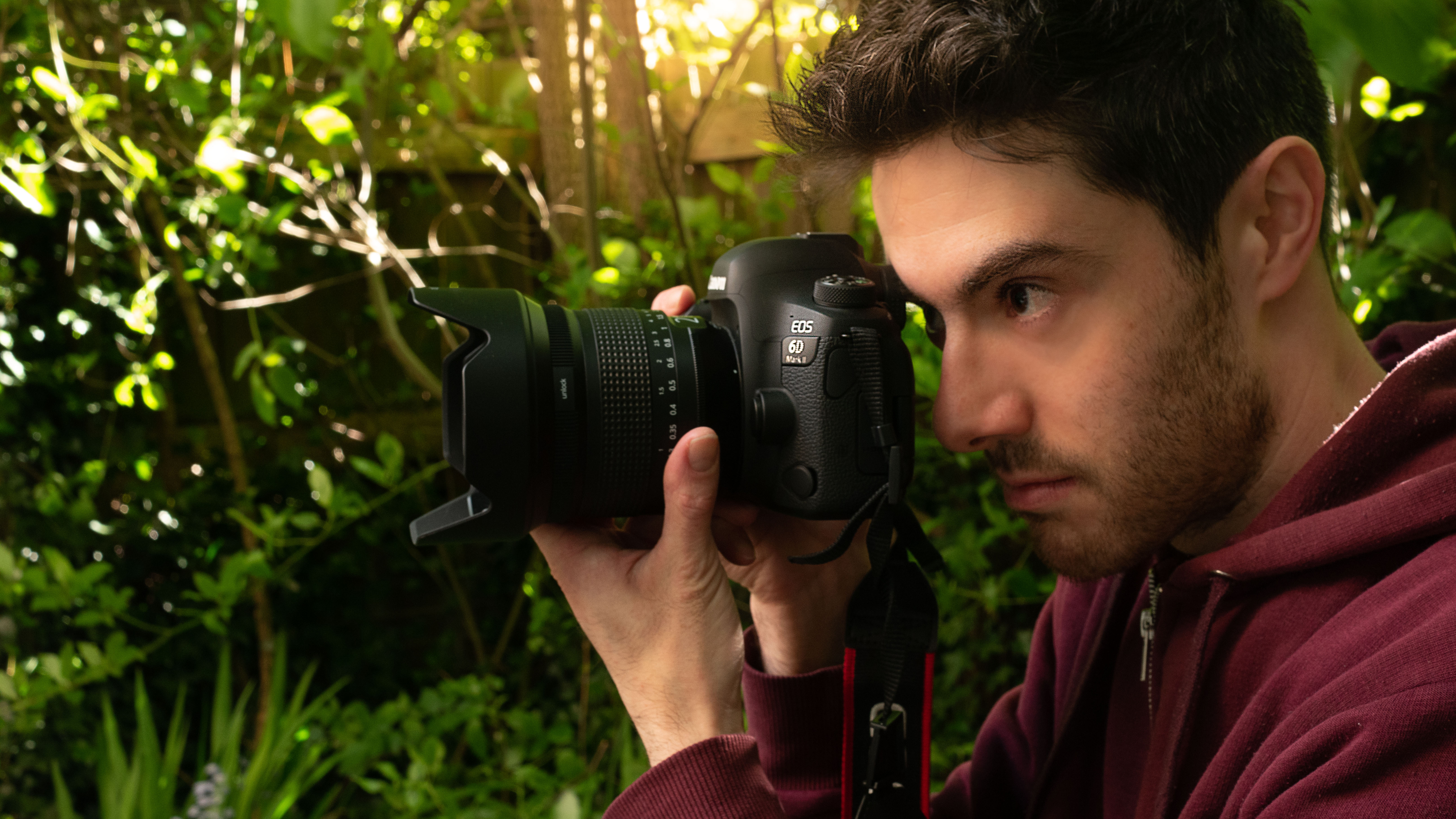 A man holding a Canon EOS 6D Mark II camera and an Irix lens, in an outdoor environment with sunlight filtering through vegetation in the background