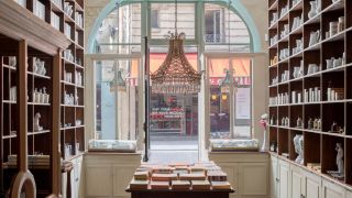 The interior of an old-school perfumery sculpted in wood and glass, with cabinets filled with ceramics, candles, and books, features a central chandelier overlooking an empty road.