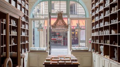 The interior of an old-school perfumery sculpted in wood and glass, with cabinets filled with ceramics, candles, and books, features a central chandelier overlooking an empty road.