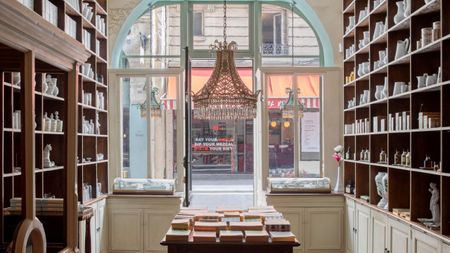 The interior of an old-school perfumery sculpted in wood and glass, with cabinets filled with ceramics, candles, and books, features a central chandelier overlooking an empty road.