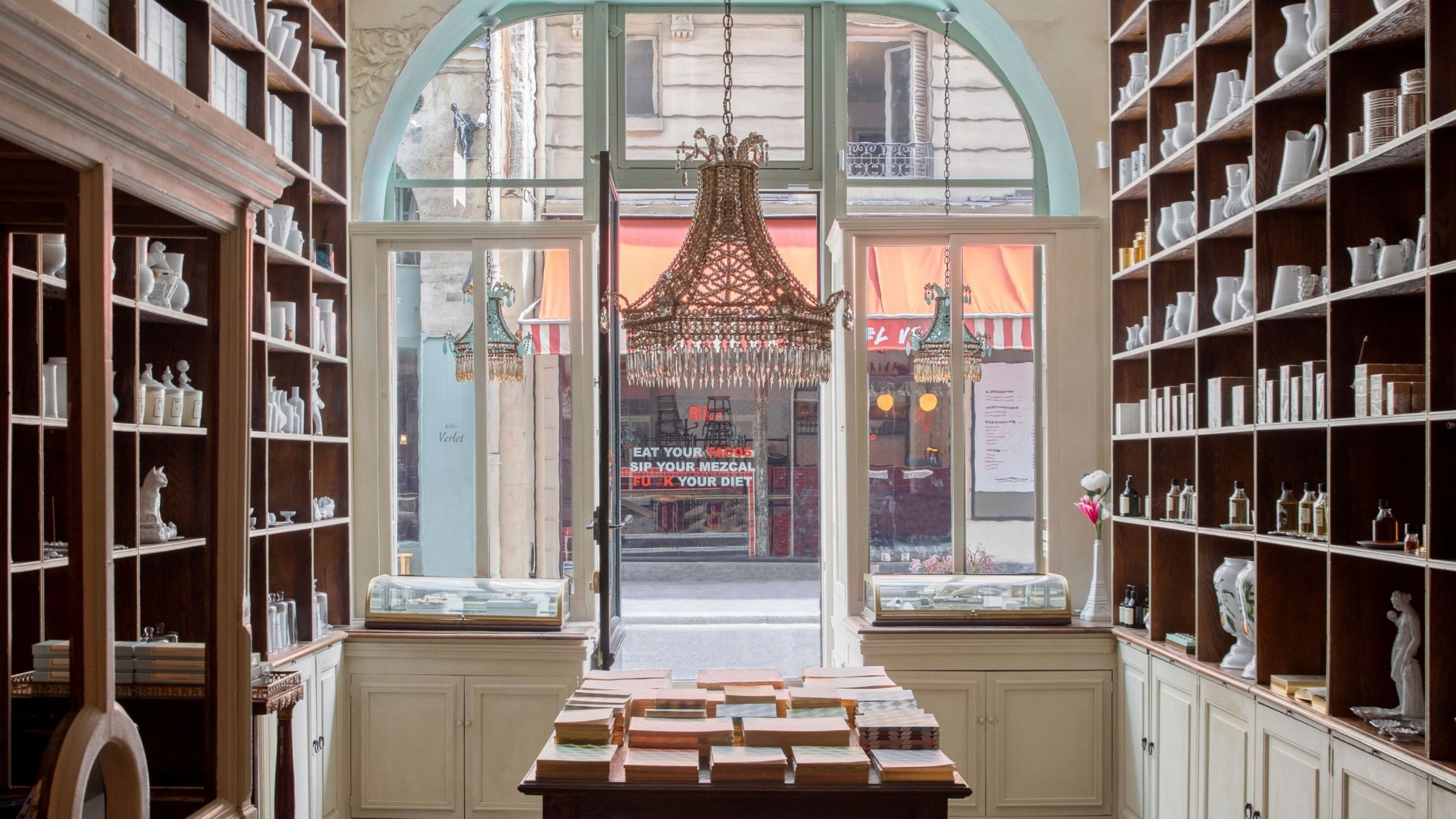 The interior of an old-school perfumery sculpted in wood and glass, with cabinets filled with ceramics, candles, and books, features a central chandelier overlooking an empty road.