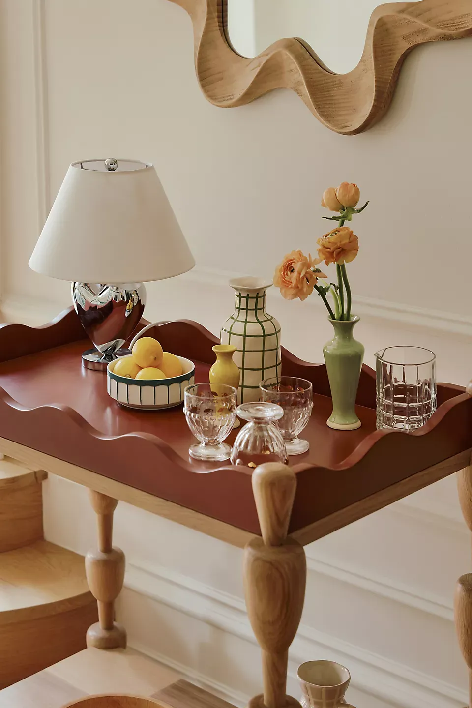 a bar cart from the anthropologie x lala reimagined collection styled with glassware, vases, a chrome lamp and bud vase, with a squiggly timber mirror hung above