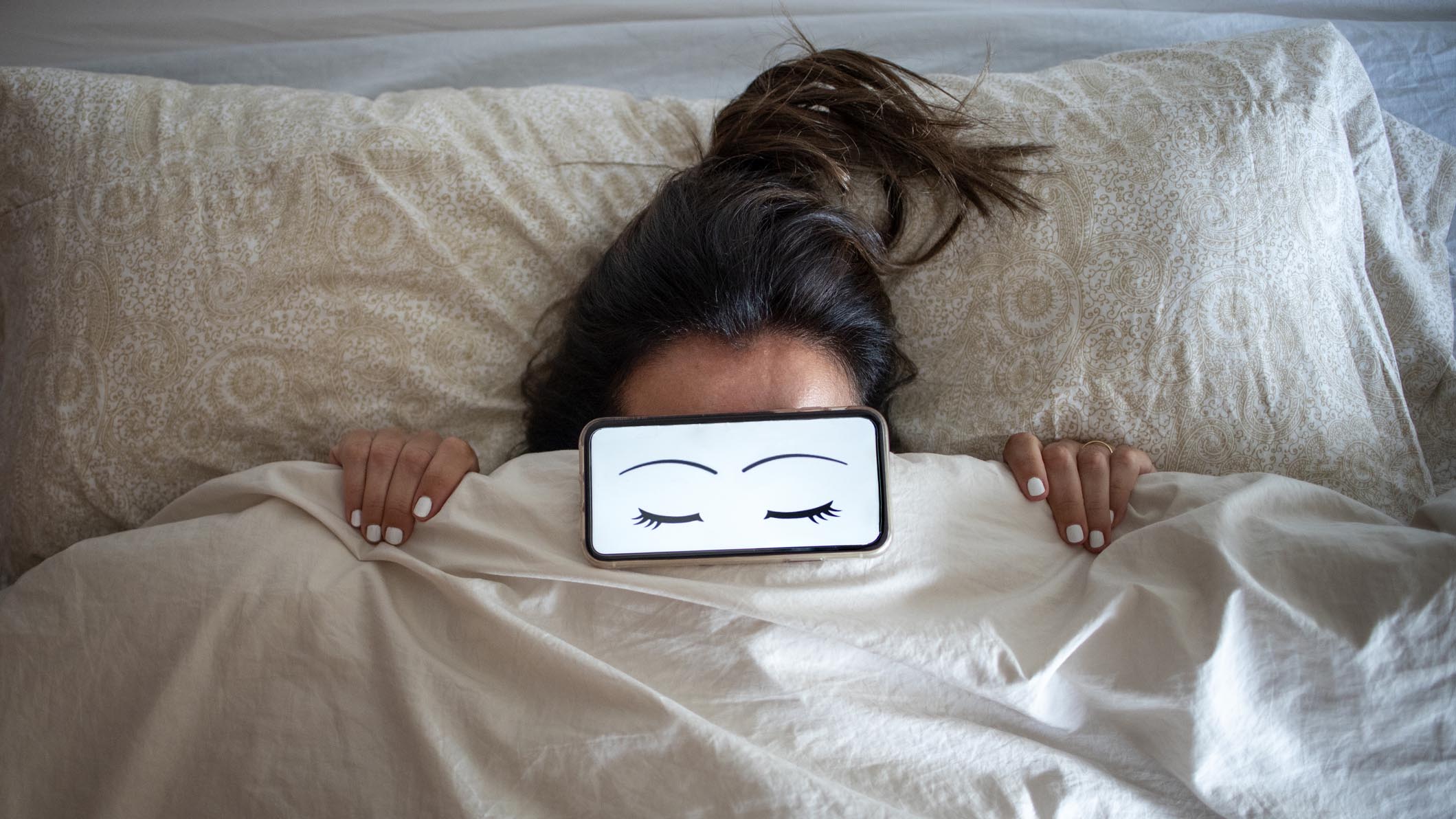 A woman lies in bed with a phone covering her eyes