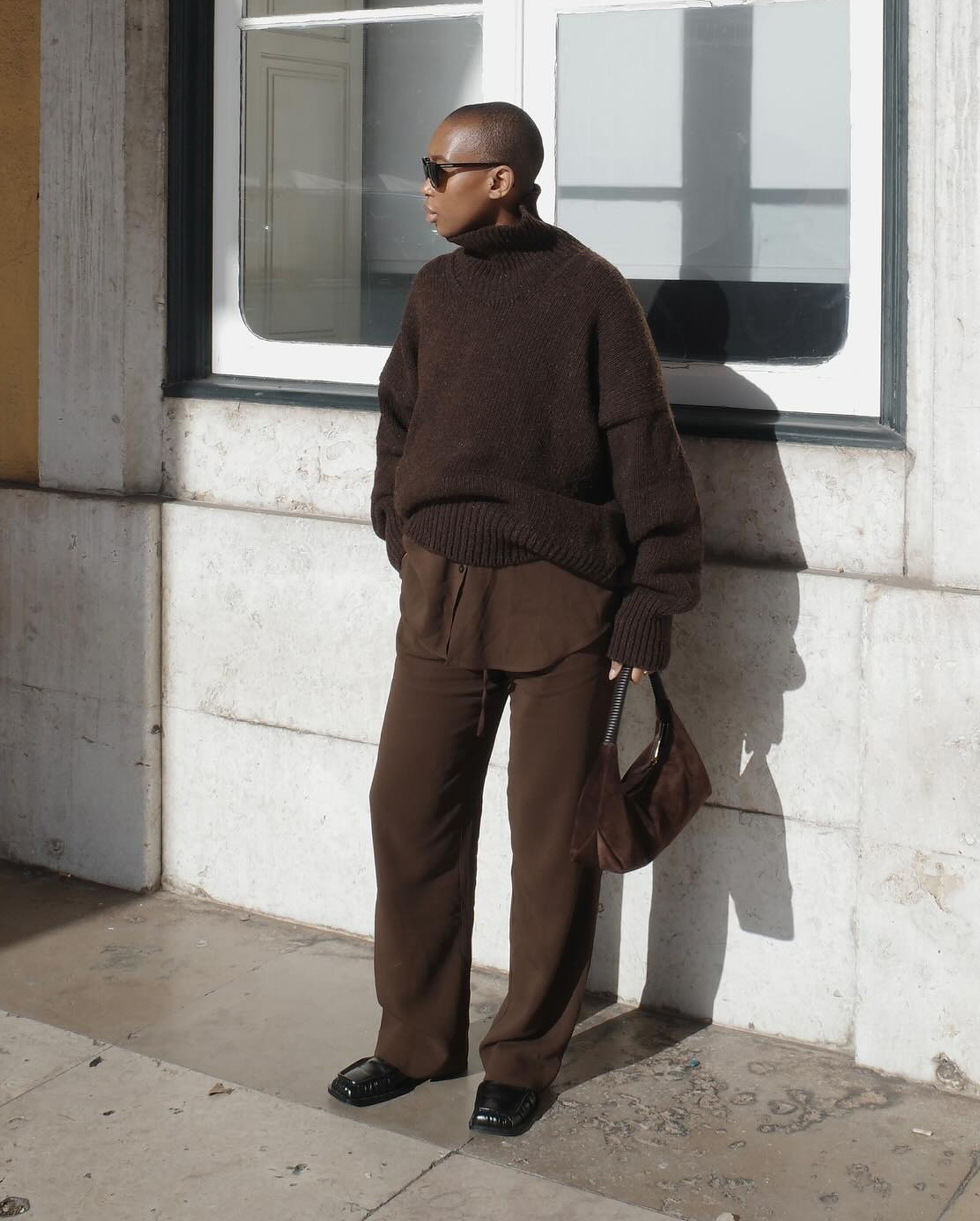 London style influencer Marisa Martins poses in a chic maternity outfit wearing sunglasses an oversize brown sweater layered over a brown button-down shirt, a brown suede shoulder bag, brown drawstring pants, and black square-tow ruched loafers