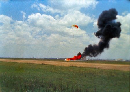 A man parachutes from a fiery crash in a field far away. 