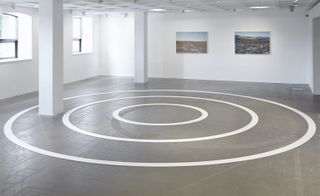 A gallery with white wall featuring wall art and 3 white cirlcles (in different circles) in the center of the room on grey floor