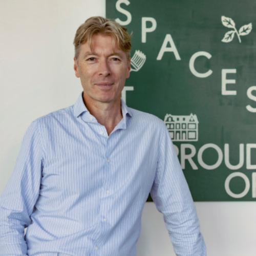 A man in a light blue button-down shirt standing in front of a white wall that has a green poster hanging on it.