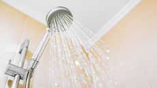 A view looking up at a running metal shower head, attached to a metal bar on a tan tiled bathroom with tiled walls and a white ceiling. 