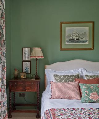 Traditional green guest bedroom with cushions on bed, mid century side table, lamp and walled art depicting a ship and other ones showing painted florals