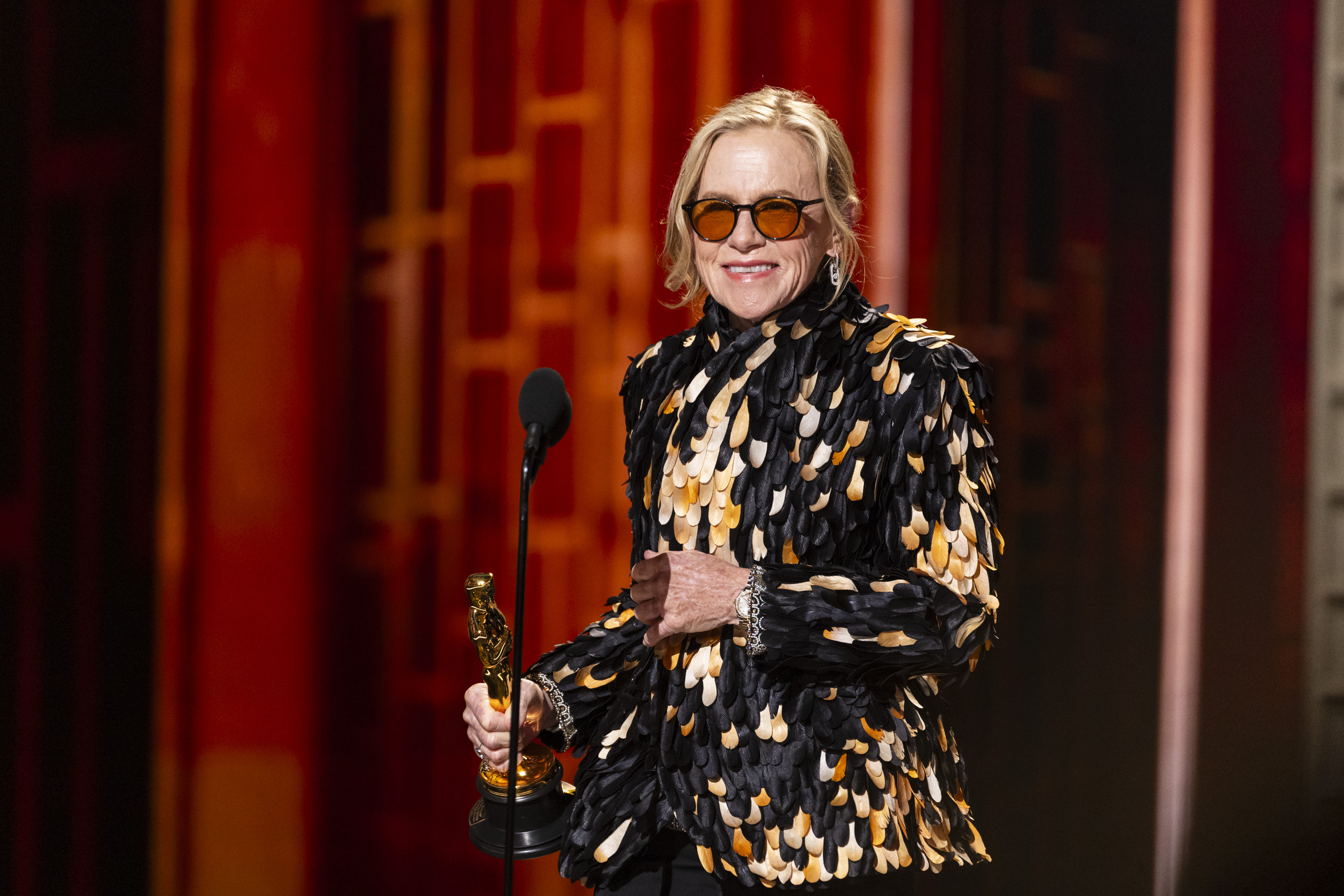 Amy Madigan wears a feathered top and sunglasses and smiles as she accepts the oscar for best supporting actress at the 2026 oscars
