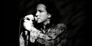 Eddie Vedder with a plaid shirt on singing into a microphone in black and white.