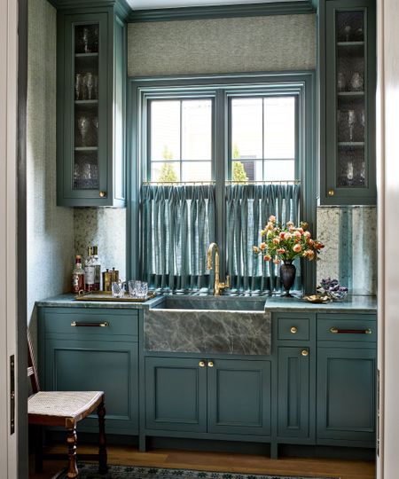 Blue home bar with blue painted cabinetry, marble sink and countertops, a large window at the centre with a blue cafe curtain, two cabinets of glassware and a wooden chair 