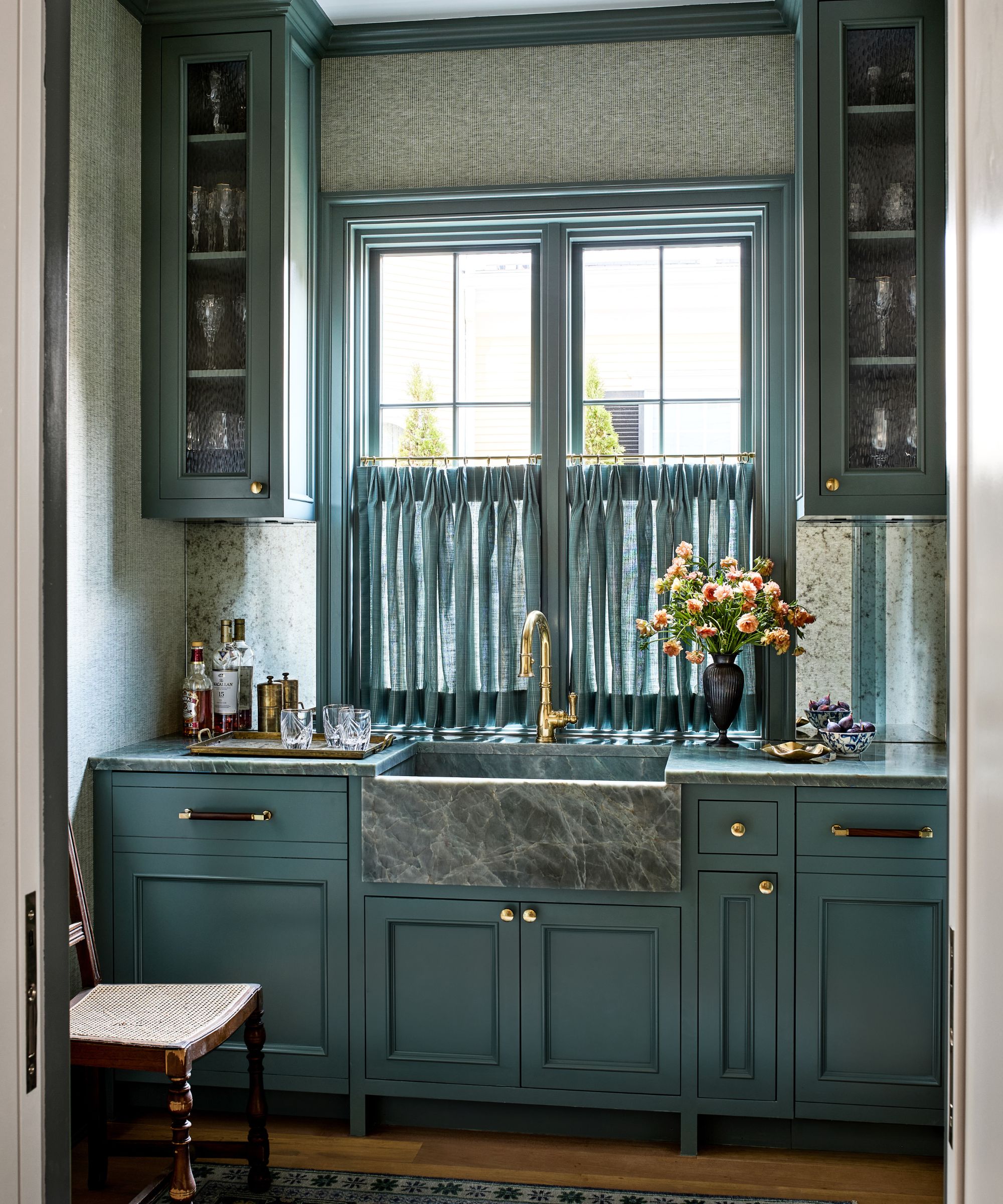 Blue home bar with blue painted cabinetry, marble sink and countertops, a large window at the centre with a blue cafe curtain, two cabinets of glassware and a wooden chair