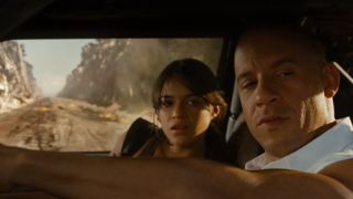 Vin Diesel driving a car with Michelle Rodriguez in the passenger seat and an explosion behind them.