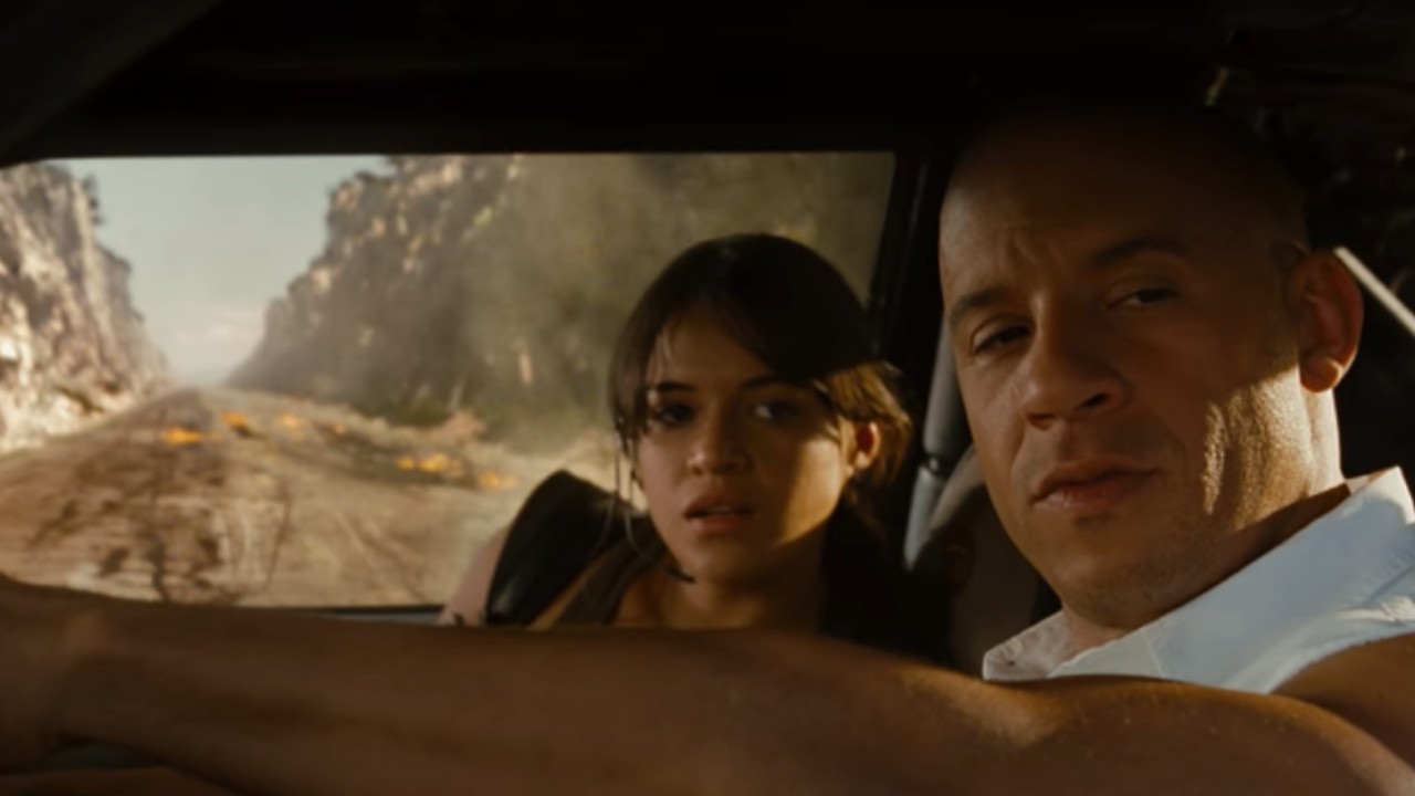 Vin Diesel conduciendo un auto con Michelle Rodriguez en el asiento del pasajero y una explosión detrás de ellos.