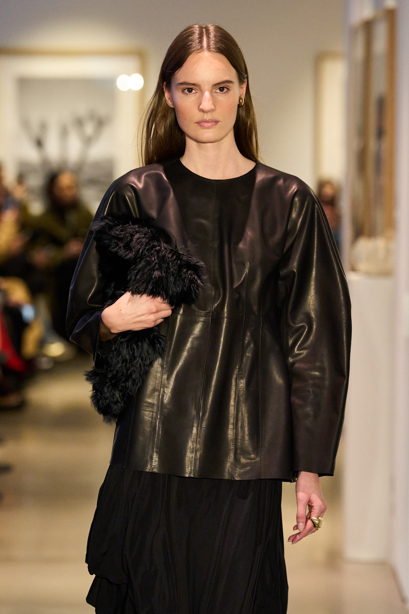 Fur accessory trend is shown in Ashlyn's fall-winter 2025 collection in a close photo of a model walking down the runway wearing a curved leather top with a black maxi skirt and a black furry clutch bag