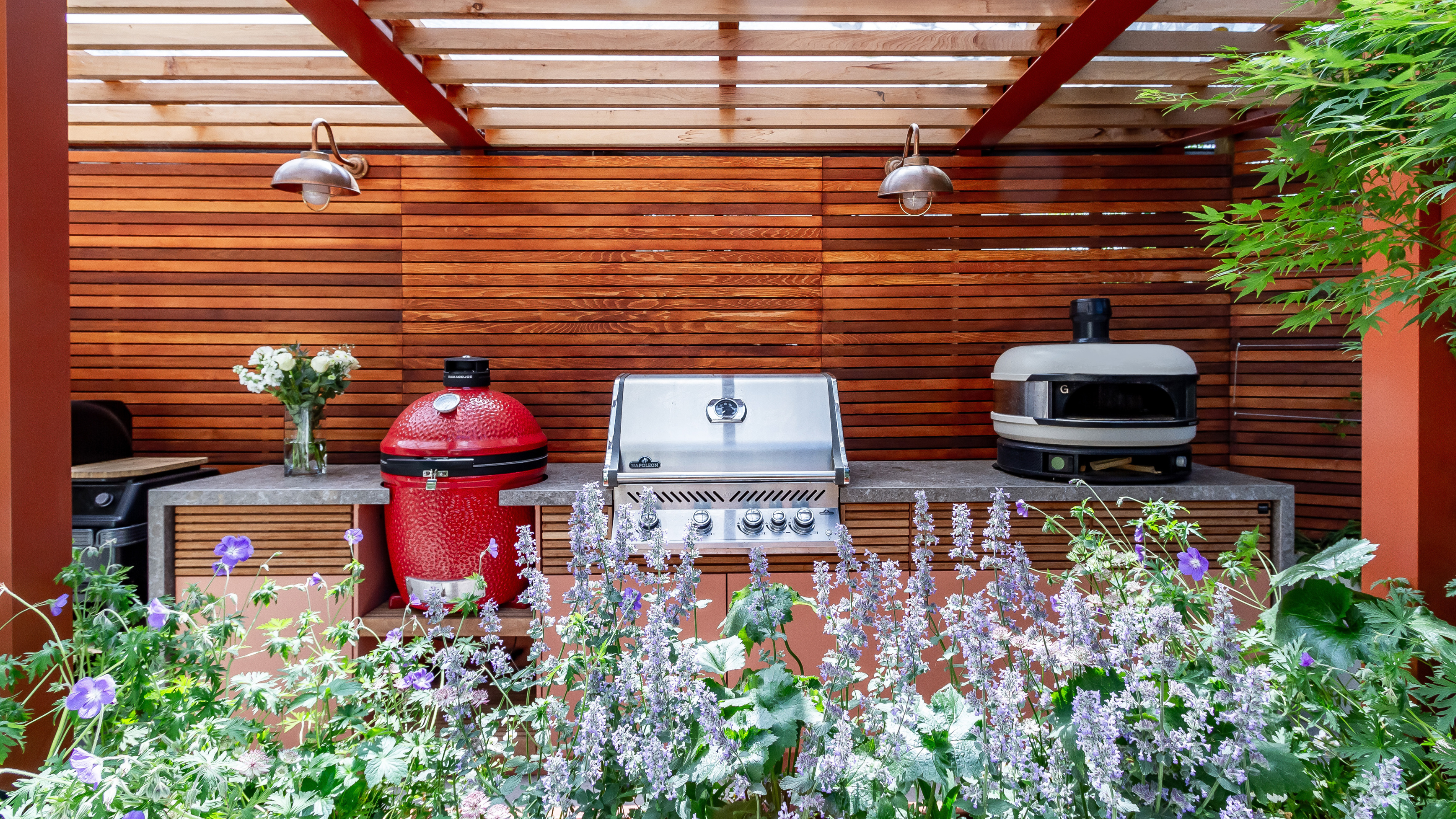a wooden pergola with an outdoor kitchen with three different cooking appliances including a built-in grill, green egg and gozney pizza oven with plants in the foreground