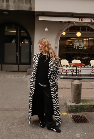 what to wear in 30-degree temperatures shown in a photo of a woman standing on the sidewalk wearing a long zebra print coat over a white t-shirt layered under a black cashmere sweater styled with black trousers and black sneakers