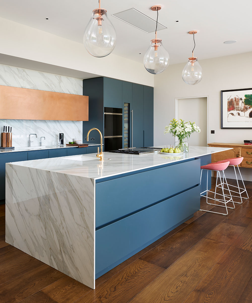 A blue painted kitchen island with marble waterfall top and pink bar stools.