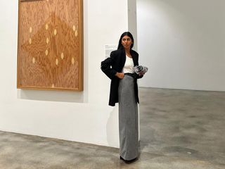 Fashion influencer Monikh Dale in an art gallery wearing a long gray skirt, a white top, and a long black blazer and carrying a clutch handbag.