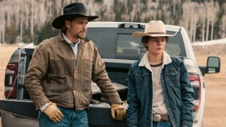 L-R): Luke Grimes as Kayce Dutton and Brecken Merrill as Tate Dutton standing behind a truck.