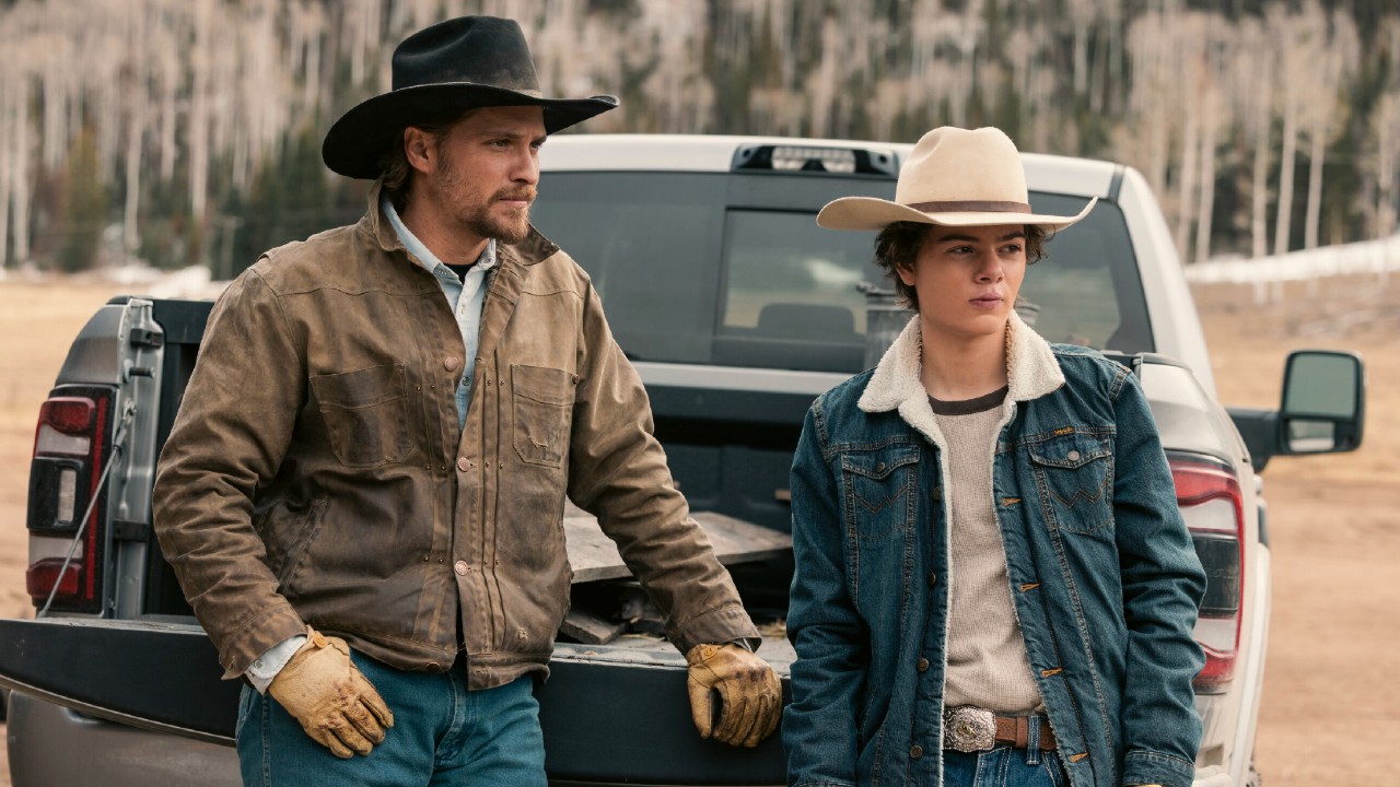 L-R): Luke Grimes as Kayce Dutton and Brecken Merrill as Tate Dutton standing behind a truck.