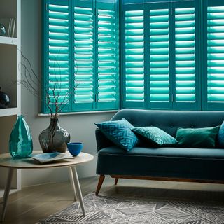 Living room window with teal velvet sofa below and windows dressed in blue shutters