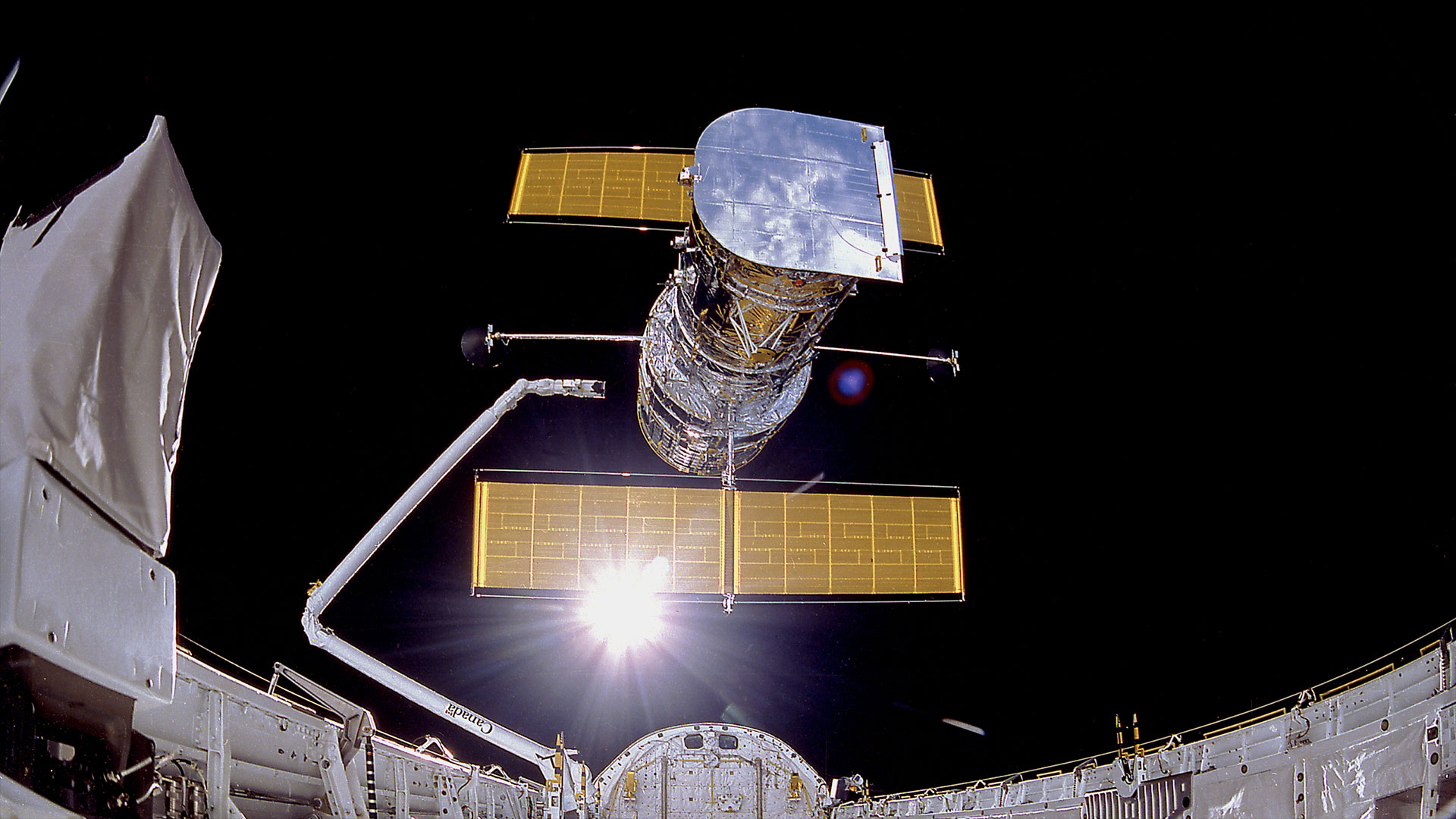 a large silver satellite with orange solar array wings is released from a robotic arm into the glare of the sun