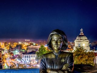 A bronze statue of Star Trek Voyager's Captain Janeway stands with crossed arms in front of a small downtown area at light, the streets light with many colors of nightlife, and the tall dome of a limestone courthouse on the right side of the statue.