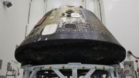 A space capsule with charred black on its bottom sits inside of a tall clean room. 