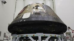 A space capsule with charred black on its bottom sits inside of a tall clean room. 