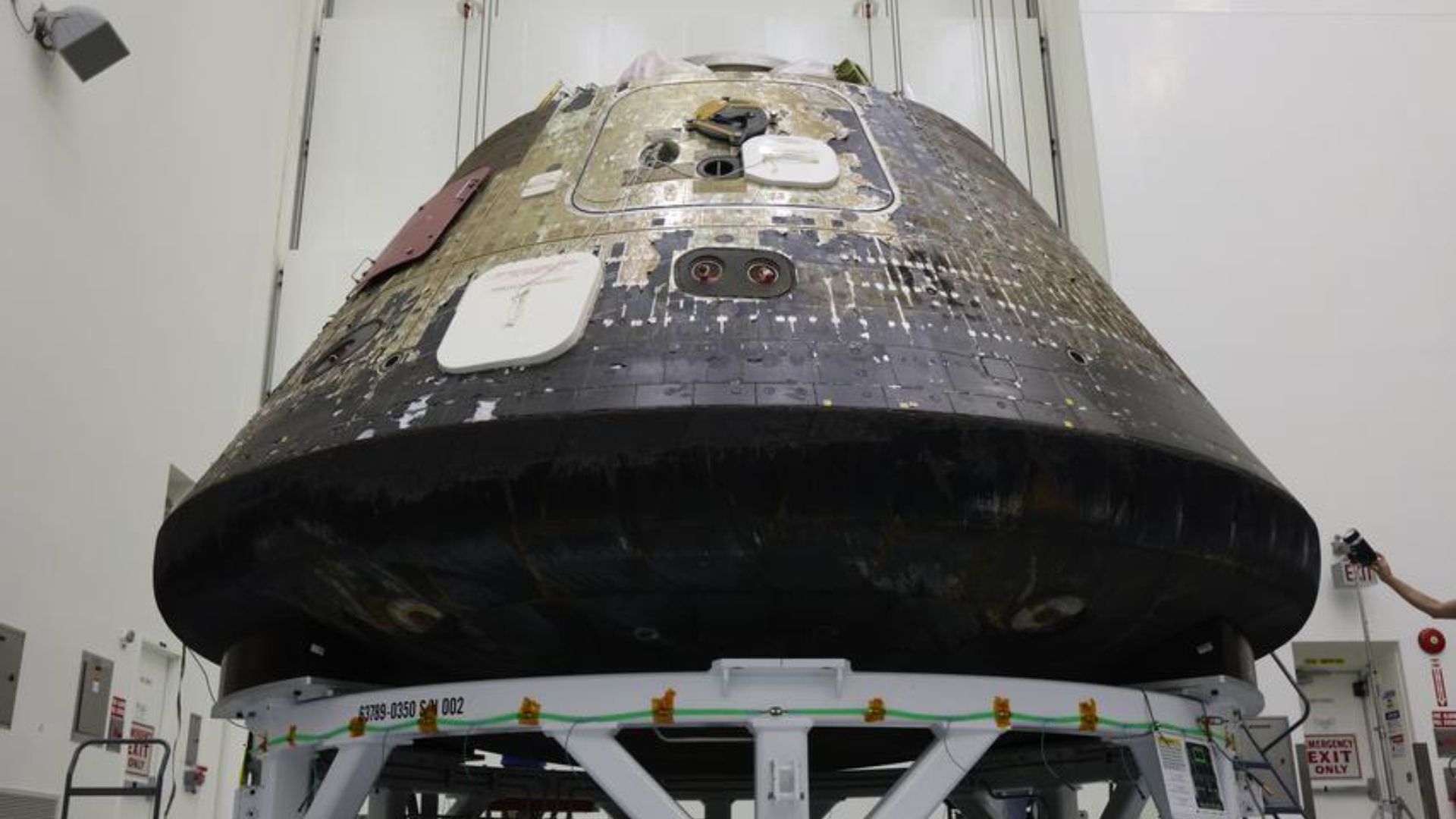 A space capsule with charred black on its bottom sits inside of a tall clean room. 