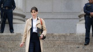 Lizzy Caplan as Alex Mullen walking down the stairs of a government building wearing a dark suit, white shirt and beige trench coat while police guard the outside of the building.