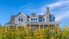 External shot of large house with gray exterior brickwork. It has peaked windows and a wraparound porch, set against a bright blue sky and yellow wildflowers in foreground