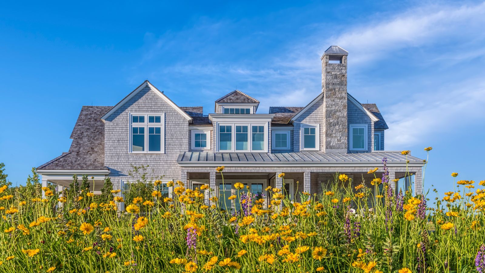 External shot of large house with gray exterior brickwork. It has peaked windows and a wraparound porch, set against a bright blue sky and yellow wildflowers in foreground