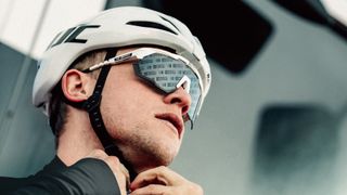 A white man with some stubble and a black jersey fastens his white helmet. He is wearing a large pair of sunglasses that have white frames and lenses monogrammed with small logos