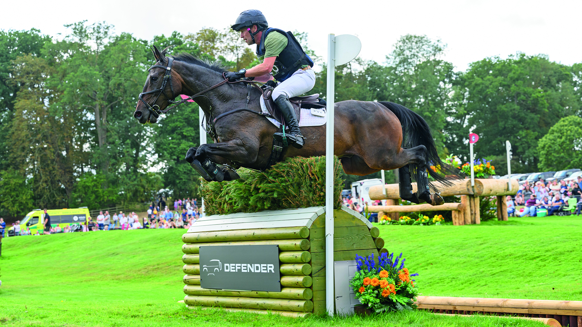 Simon Grieve, a rider in a green T-shirt and black body protector, rides bay horse Autograf over a fence with a hedge top at Burghley 2025