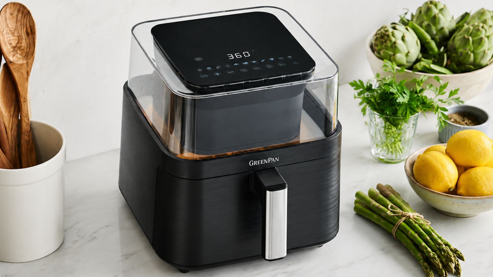 The GreenPan Bistro 360 Clear View Air Fryer on a marble kitchen counter beside a crock of wooden utensils and some fresh spring veg. 