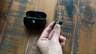 The Beats Studio Buds in black pictured on a wooden surface and someone is holding the left bud