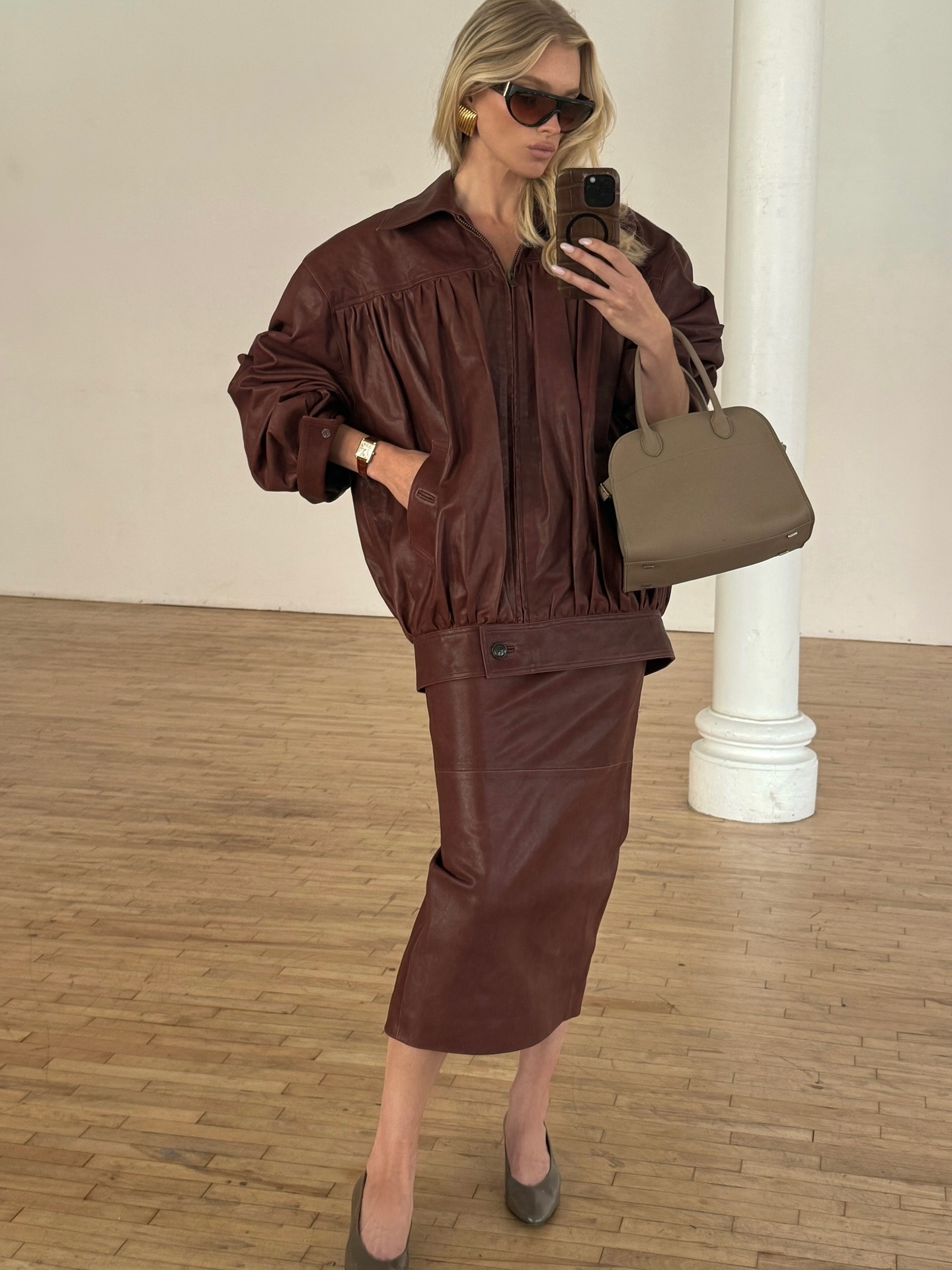Model @hoskelsa takes a mirror selfie wearing a brown leather skirt with a brown leather jacket a mid heel pumps