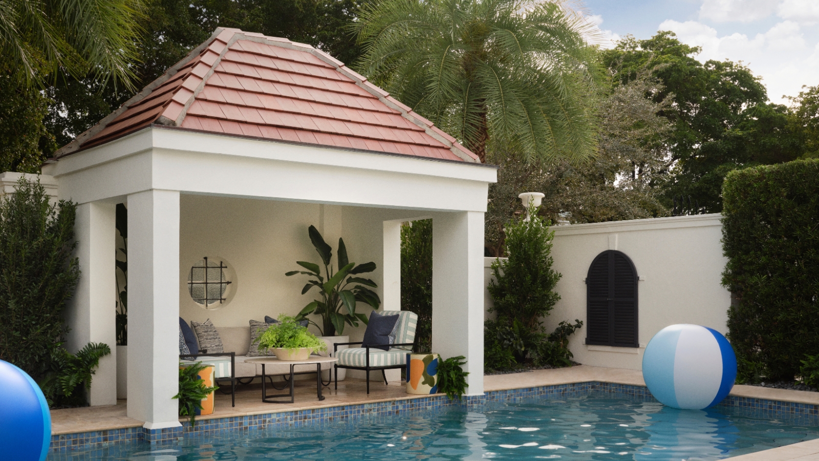 Swimming pool framed by tropical planting with a white open-sided gazebo, striped seating and terracotta-tiled roof.