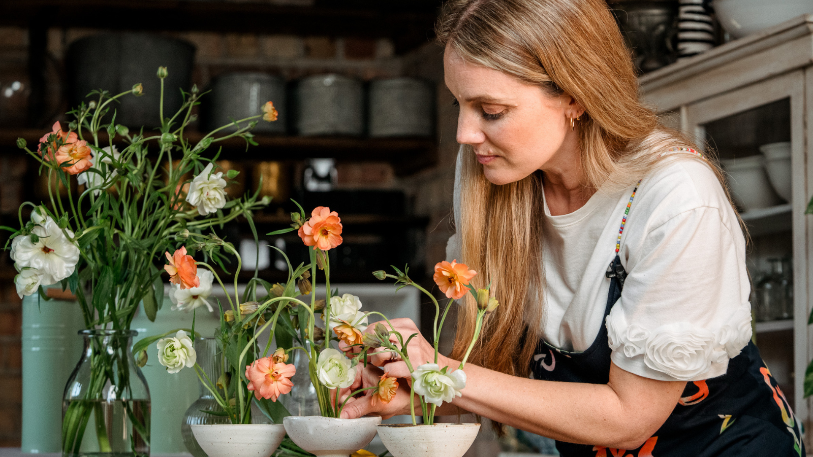 Woman creating a floral design with peach butterfly ranunculus and uva vulpis fritillaries in three ceramic bowls
