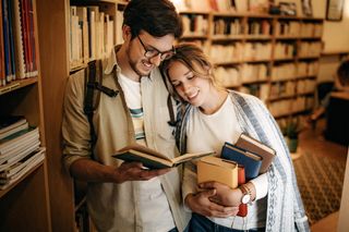 A man and woman in a book shop holding a couple of books, while cosying up together.