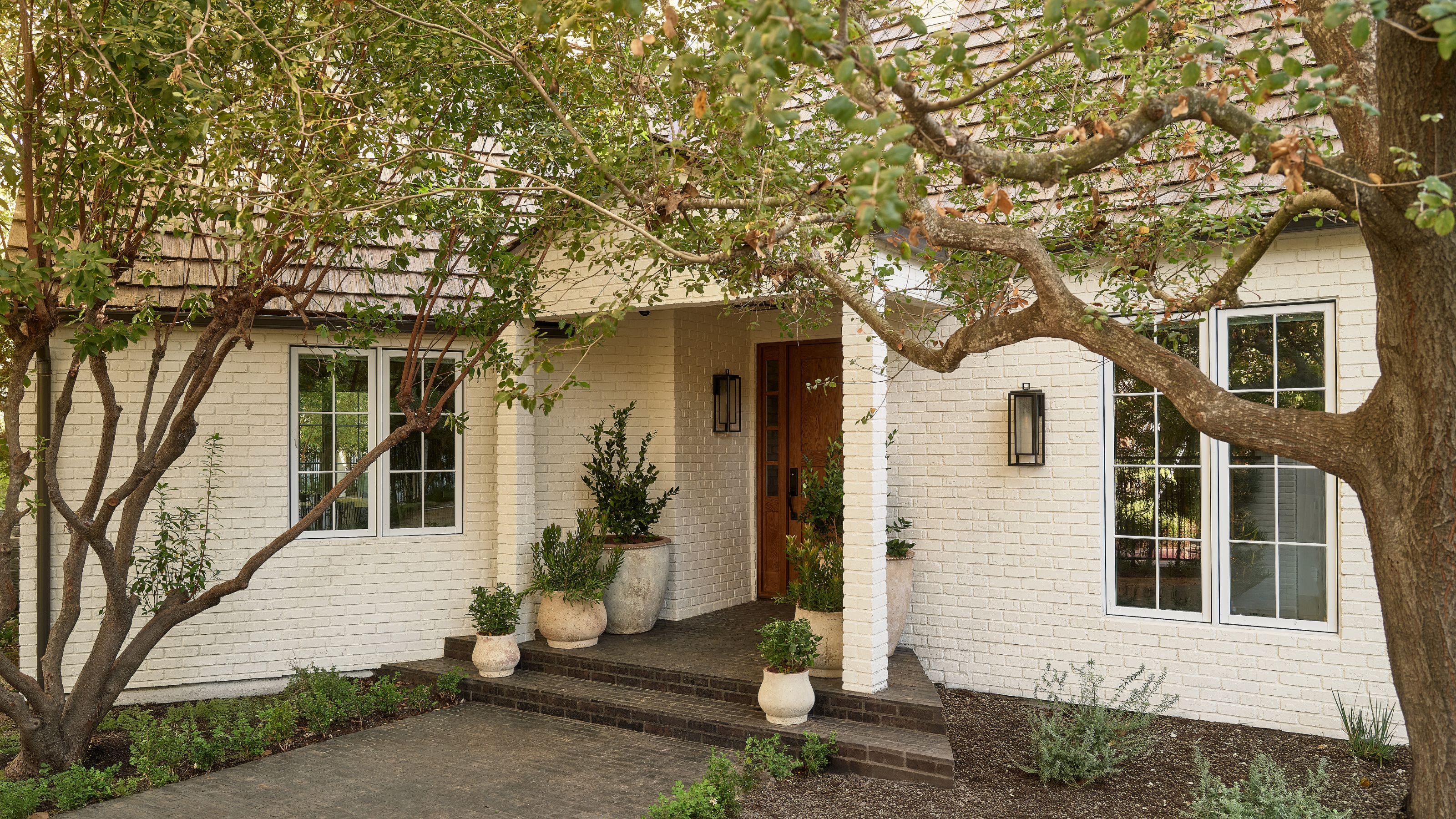 a white painted brick house with trees and potted plants on the front step