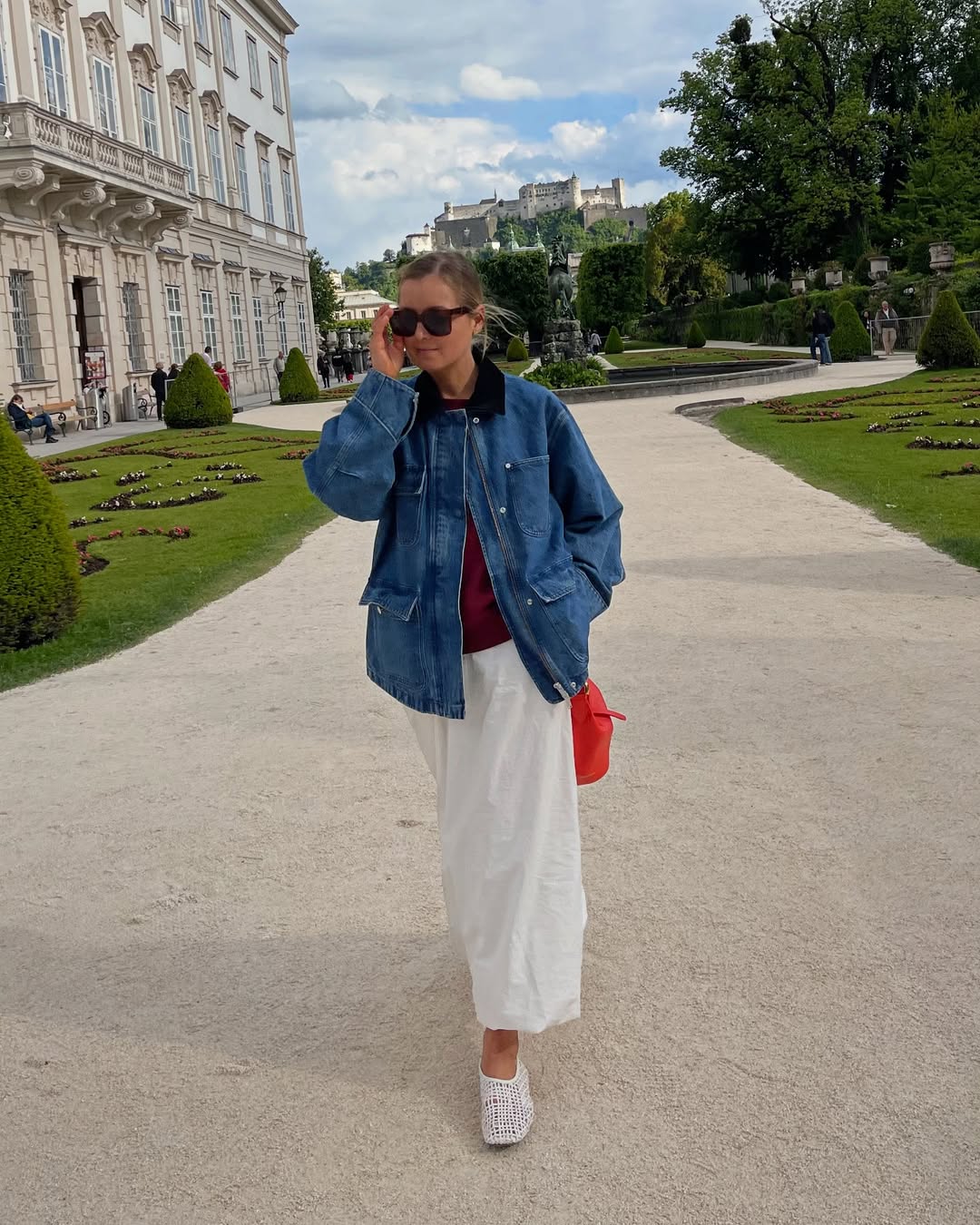 @annaaborisovna wears a denim jacket, burgundy shirt, white midi skirt, white woven flats and a bright red bag