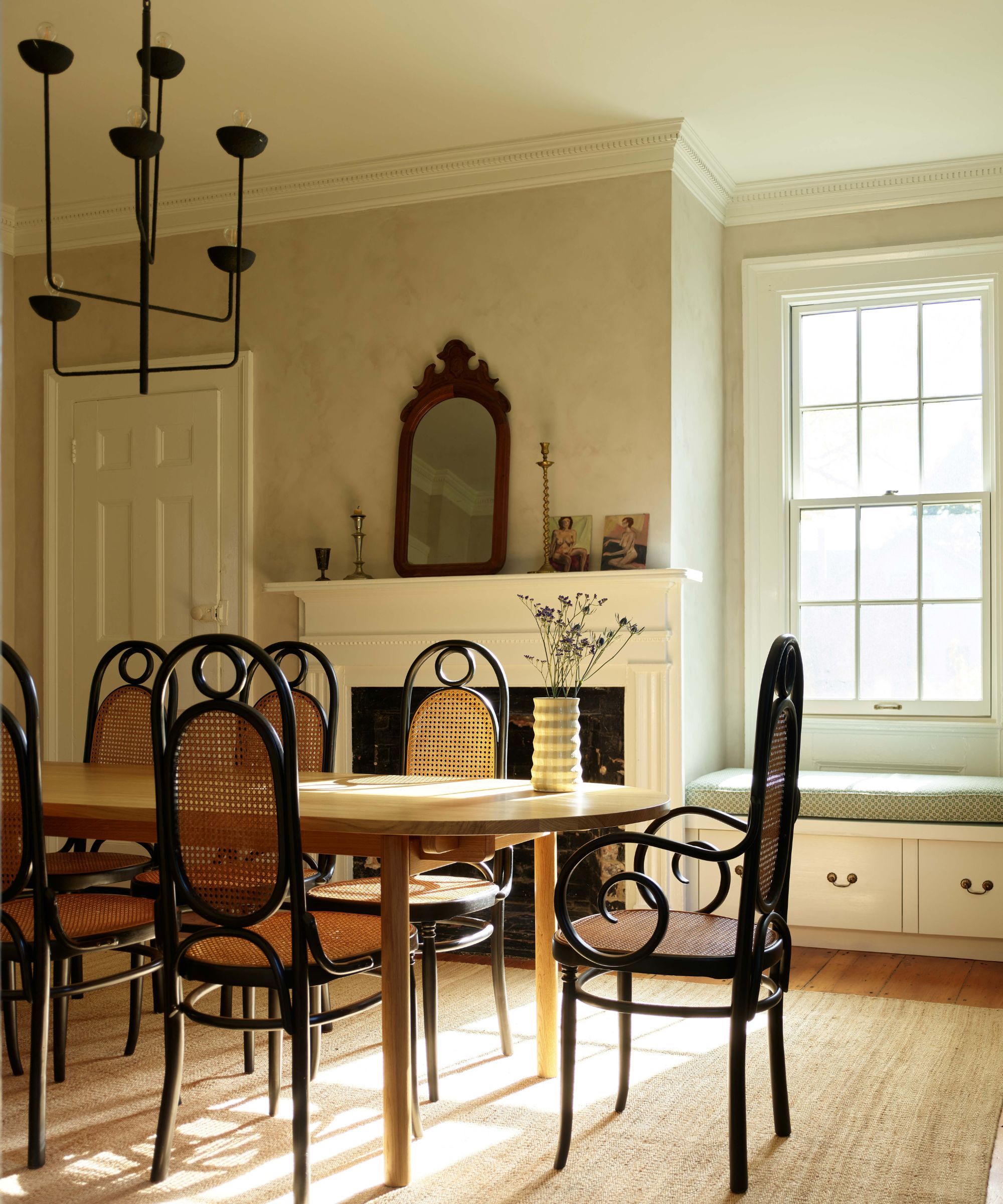 Dining room with a long oval table, several black cane-back chairs, a fireplace with a wood-framed mirror