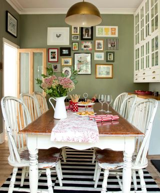 A green dining room with a gallery wall of square prints, a gold curved pendant light, a wooden dining table with a table runner, decor, white chairs and edges, and a black and white striped rug