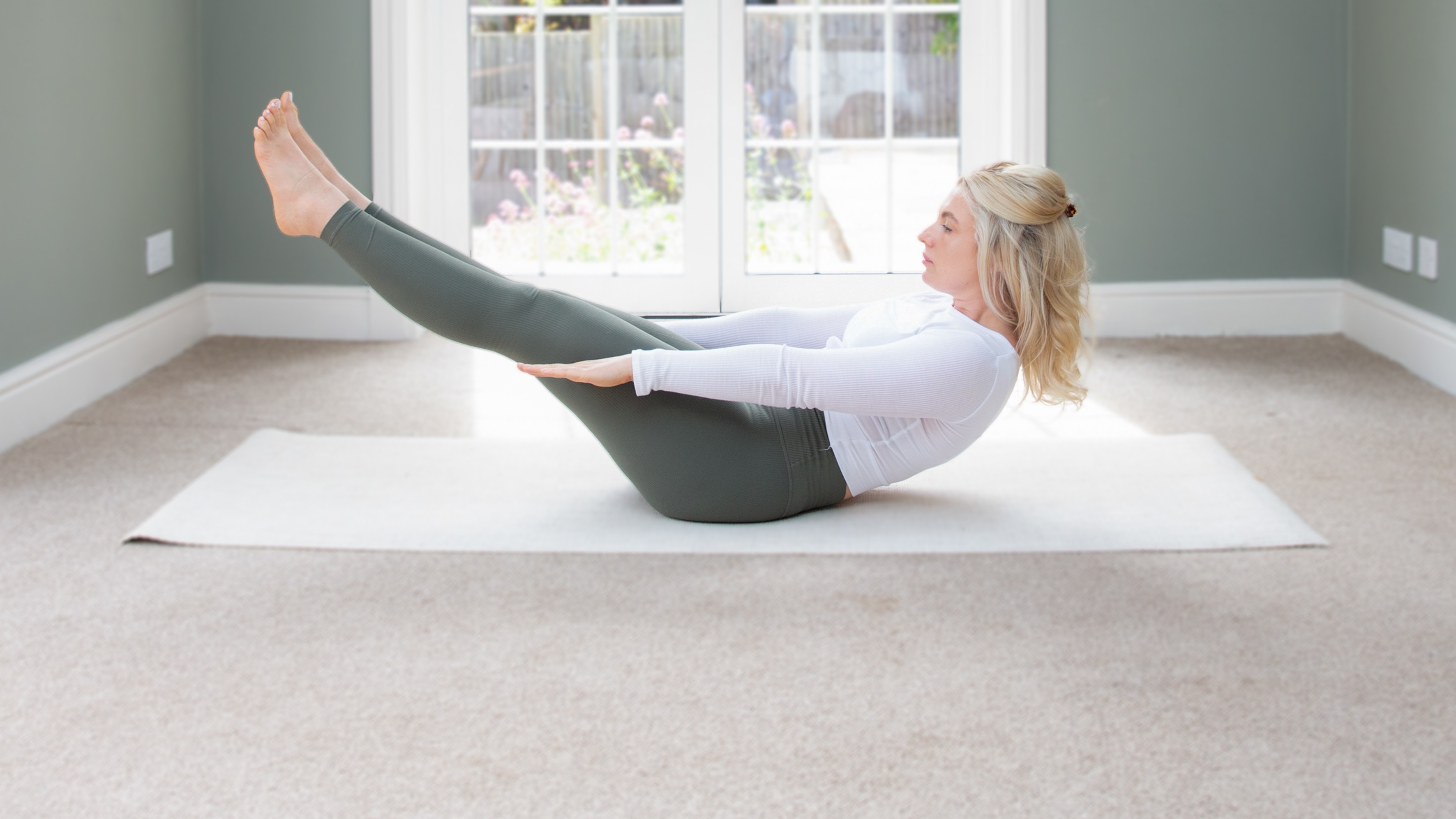 Michelle D'Onofrio demonstrates the Hundred Pilates move on an exercise mat. She is seated, with her back slightly elevated from the floor, arms straight at her sides, and legs lifted in the air.