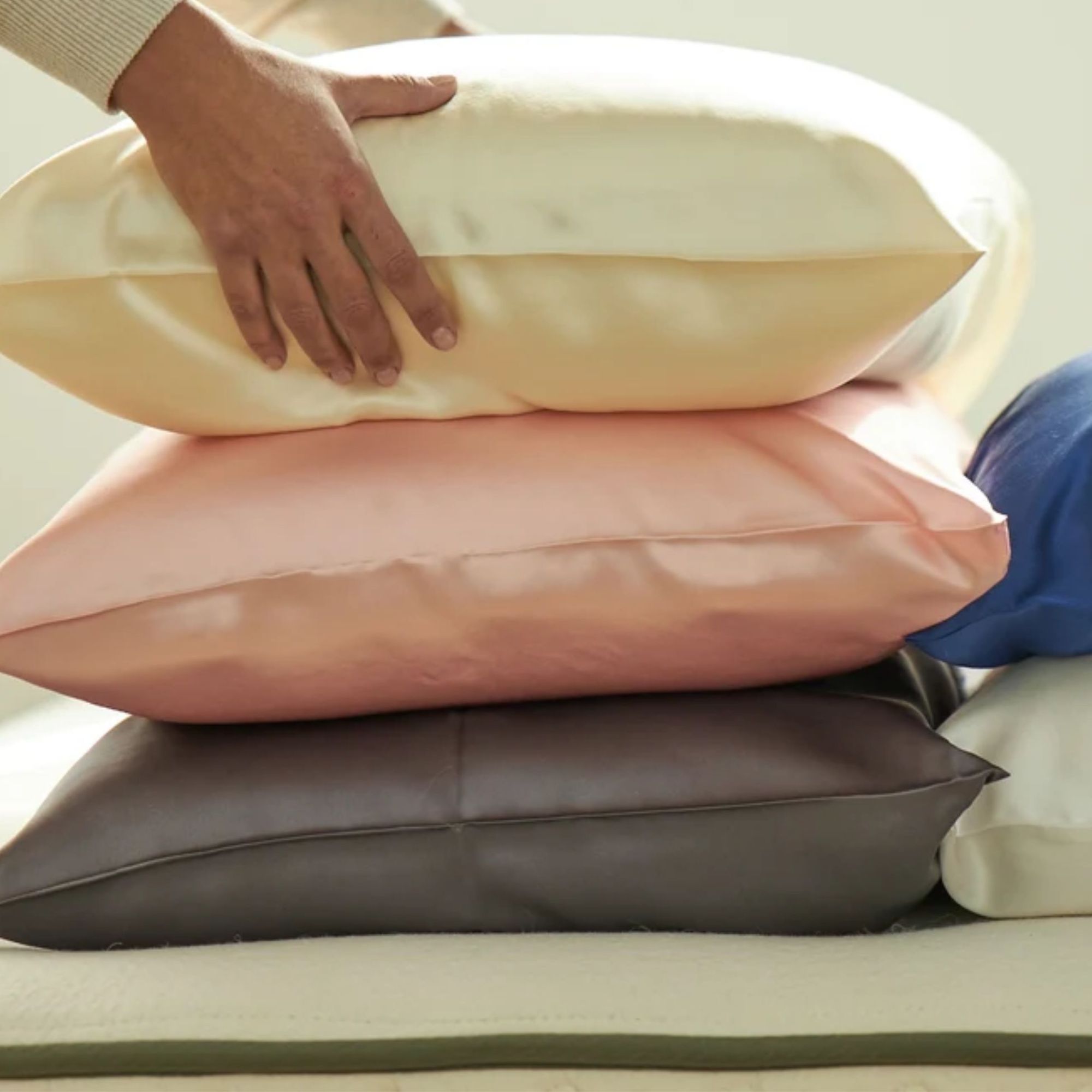 Avocado silk pillowcases in white, pink, and dark grey, stacked up on a mattress with hands holding the top one